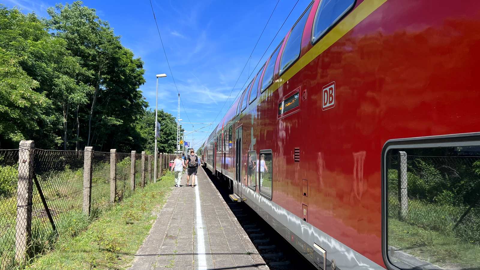 Roter Doppelstockzug der DB am einzigen Bahnsteig des Bahnhofs Raddusch, daneben Fahrgäste auf dem Bahnsteig, ein Maschendrahtzaun und Bäume unter blauem Himmel